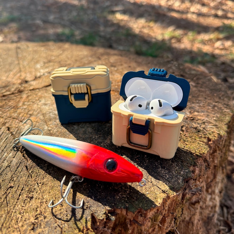 Puede incluir: Un se&ntilde;uelo de pesca rojo, blanco e iridiscente con anzuelos triples descansa sobre un toc&oacute;n de &aacute;rbol junto a dos estuches en miniatura con forma de nevera. Un estuche est&aacute; abierto, revelando auriculares blancos. Los estuches son azules y beige.