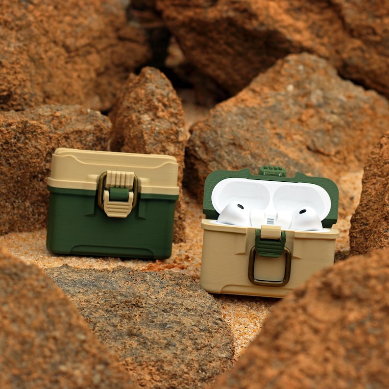 May include: Two-tone protective cases for wireless earbuds. One case is closed, the other is open, revealing white earbuds. The cases are olive green and tan, with a latch closure. The background is a sandy, rocky terrain.