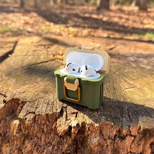 May include: A green and tan protective case for wireless earbuds. The case is open and the earbuds are inside. The case has a latch and a handle.