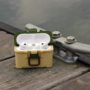 May include: A tan and olive green protective case for wireless earbuds, open to reveal white earbuds. The case has a hinged lid and a clasp closure. The background shows a wooden dock and a metal cleat.