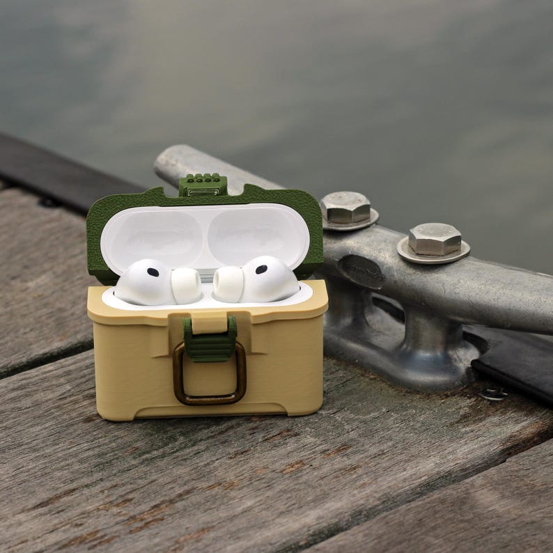 May include: A tan and olive green protective case for wireless earbuds, open to reveal white earbuds. The case has a hinged lid and a clasp closure. The background shows a wooden dock and a metal cleat.