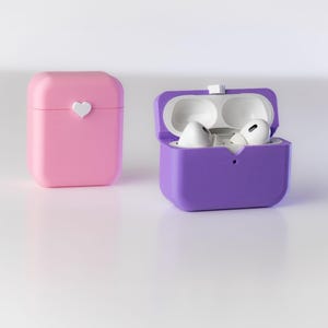 Pu&ograve; includere: Due custodie protettive per auricolari wireless. Una custodia &egrave; rosa con un design a forma di cuore bianco. L'altra custodia &egrave; viola ed &egrave; aperta, rivelando due auricolari bianchi all'interno.