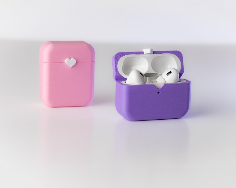 Pu&ograve; includere: Due custodie protettive per auricolari wireless. Una custodia &egrave; rosa con un design a forma di cuore bianco. L'altra custodia &egrave; viola ed &egrave; aperta, rivelando due auricolari bianchi all'interno.
