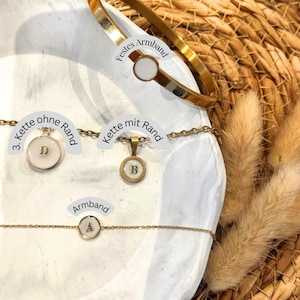May include: A selection of gold-coloured jewellery, including a bangle and necklaces with initial pendants. The bracelet and pendants have a white enamel background. The image includes the text "Festes Armband", "Kette ohne Rand", and "Kette mit Rand".