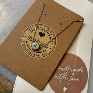 May include: A gold chain necklace with a pendant displaying the letter "A" on a white disc. The necklace is presented on a brown card with a "handmade with love" sticker and a "thank you" sticker.