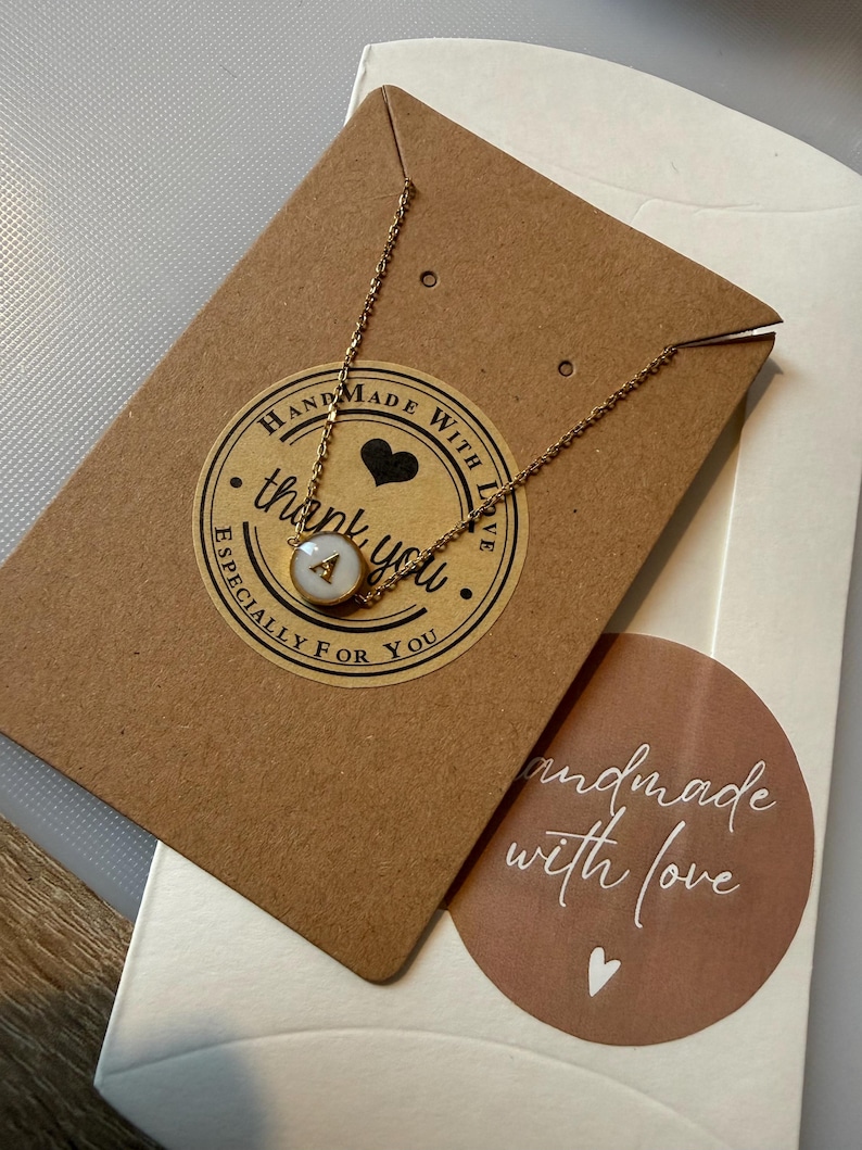 May include: A gold chain necklace with a pendant displaying the letter "A" on a white disc. The necklace is presented on a brown card with a "handmade with love" sticker and a "thank you" sticker.