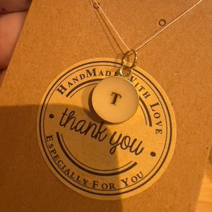 May include: A gold necklace with a circular pendant bearing the letter "T". The pendant is cream-coloured with a black letter. The necklace is presented on a brown card with a "thank you" sticker.