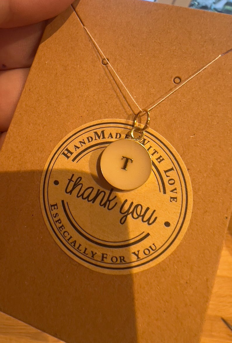 May include: A gold necklace with a circular pendant bearing the letter "T". The pendant is cream-coloured with a black letter. The necklace is presented on a brown card with a "thank you" sticker.