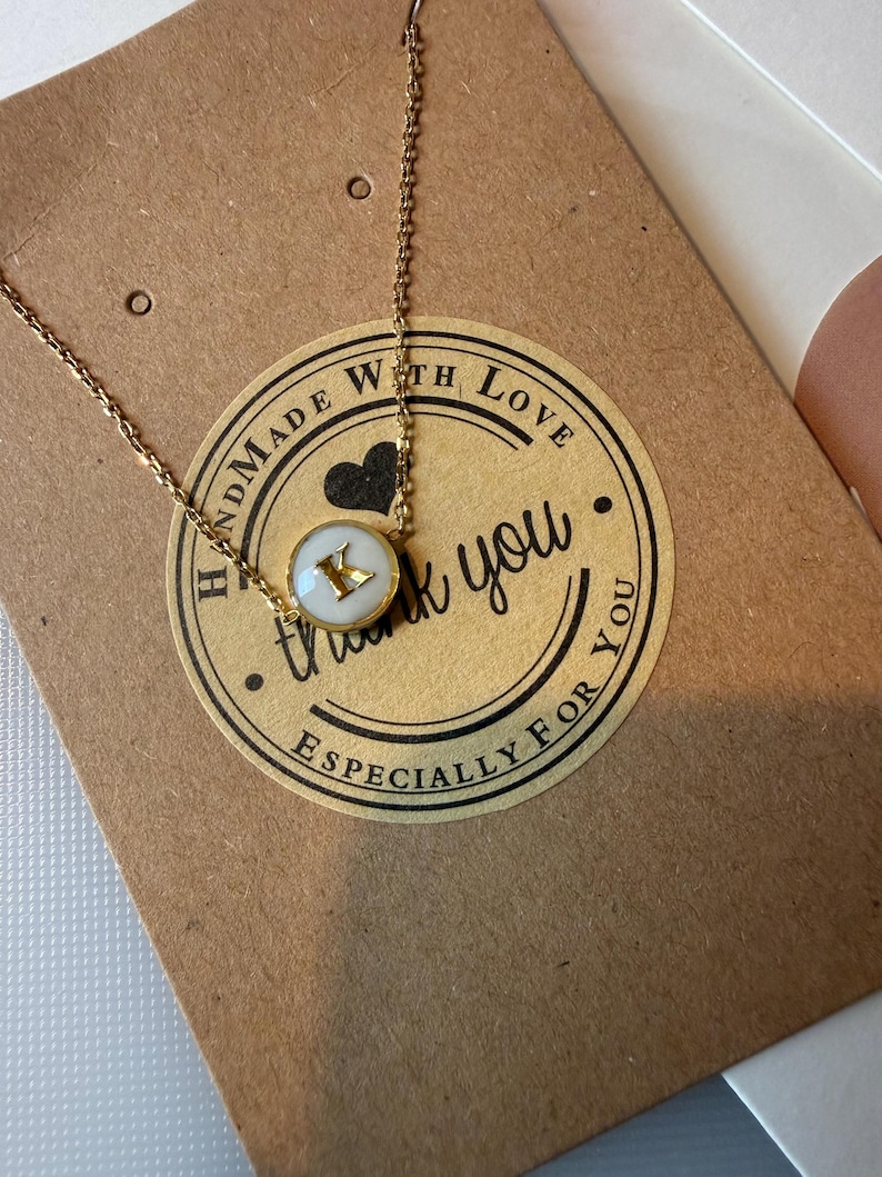May include: A gold necklace with a pendant displaying the letter "K" on a white background. The necklace rests on a brown card with a sticker that reads "Handmade with Love", "thank you", and "Especially for you".