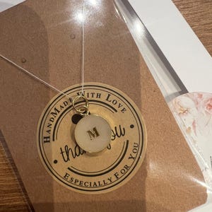 May include: A gold pendant necklace with a circular charm bearing the letter "M". The charm has a white centre and a gold edge. The pendant is suspended on a thin, clear cord. The necklace is presented on a brown card with a "Handmade with Love" sticker.