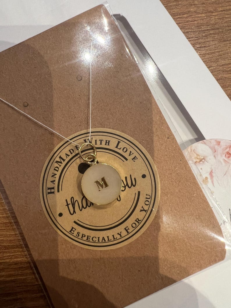 May include: A gold pendant necklace with a circular charm bearing the letter "M". The charm has a white centre and a gold edge. The pendant is suspended on a thin, clear cord. The necklace is presented on a brown card with a "Handmade with Love" sticker.