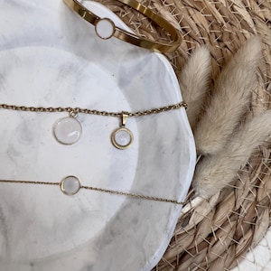 May include: A gold-coloured jewellery set comprising a bangle and two necklaces, all with circular white details. The items are presented on a white, marble-effect surface, with a woven background and dried decorative plants.