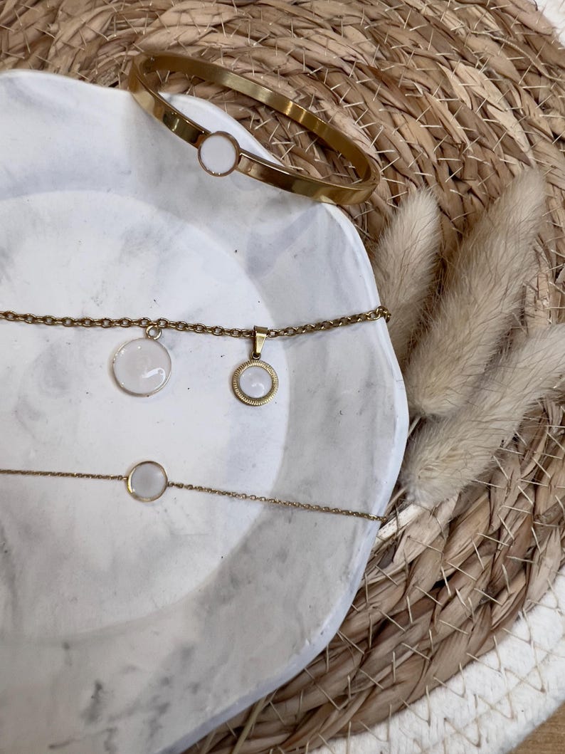May include: A gold-coloured jewellery set comprising a bangle and two necklaces, all with circular white details. The items are presented on a white, marble-effect surface, with a woven background and dried decorative plants.