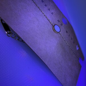 May include: A gold-coloured chain bracelet featuring a circular charm with a white centre. The bracelet is presented on a tan card with the words "WEAR ME WITH". The background is a bright blue.