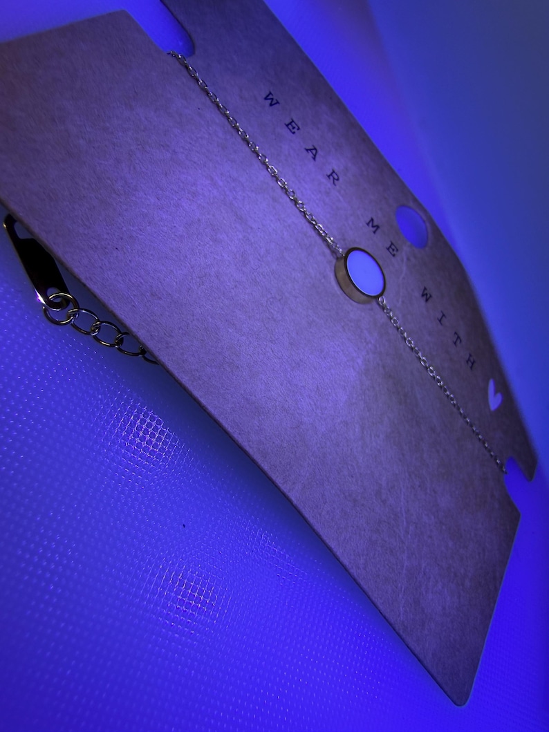 May include: A gold-coloured chain bracelet featuring a circular charm with a white centre. The bracelet is presented on a tan card with the words "WEAR ME WITH". The background is a bright blue.