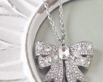May include: A silver-toned necklace featuring a bow-shaped pendant encrusted with numerous clear, sparkling rhinestones and a single rectangular faceted crystal at the center, suspended from a delicate silver-toned metal chain link necklace.