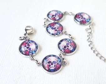 May include: A silver-toned link bracelet featuring five circular charms, each containing a graphic print of a skull surrounded by red roses and blue stained-glass patterns. The bracelet includes a lobster claw clasp and an adjustable chain link extension.