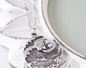 May include: A silver-toned circular pendant necklace featuring a detailed relief of a sailing ship navigating through large, textured ocean waves. The pendant hangs from a delicate silver-toned chain, showcasing a polished metallic finish.