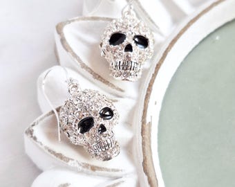 May include: A pair of silver-toned skull earrings. Each skull is encrusted with small, clear crystals, with black accents for the eyes and nose. The earrings are displayed on a white, decorative surface.