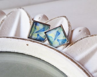 May include: A pair of square earrings with a geometric design. The earrings have a silver-colored metal frame and feature a colorful abstract pattern in shades of blue, green, and yellow. The earrings are displayed on a white surface.