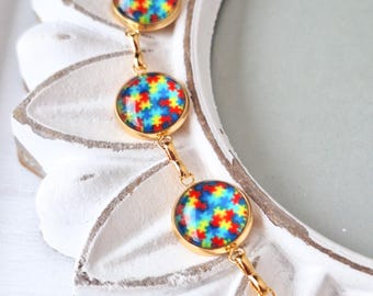 May include: A gold-toned bracelet featuring circular charms with a colorful jigsaw puzzle design. The charms are connected by delicate gold links. The bracelet is displayed against a white, ornate background.