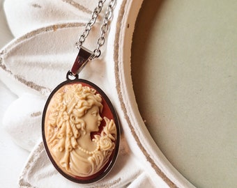 May include: A silver chain necklace with a cameo pendant featuring a woman's profile in a cream-colored cameo with a red border.