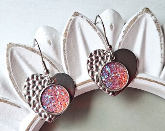 May include: A pair of silver heart-shaped earrings with a pink and iridescent glitter accent. The earrings have a textured heart shape and a round glitter accent.