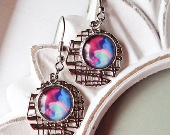 May include: A pair of silver earrings with a circular design. Each earring features a round, colorful, abstract design with shades of pink, blue, and white.