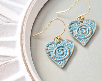 May include: A pair of heart-shaped earrings with a light blue, textured finish and gold-colored hooks. Each earring features a spiral and dot design. The earrings are displayed on a white and gray surface.