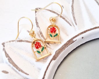 May include: Gold-tone earrings with a red rose encased in a gold cage. The rose has green leaves and a gold stem.