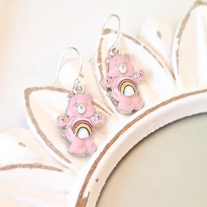 May include: A pair of silver dangle earrings featuring pink Care Bear charms with rainbow accents.