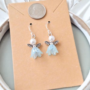 May include: A pair of dangle earrings featuring a light blue flower design with silver-tone angel wings and a white pearl bead. The earrings are displayed on a brown card with a dime.
