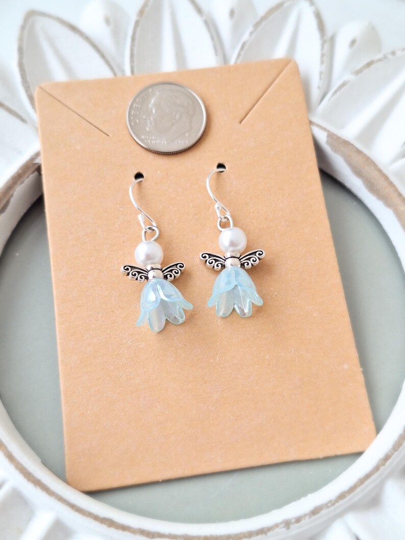 May include: A pair of dangle earrings featuring a light blue flower design with silver-tone angel wings and a white pearl bead. The earrings are displayed on a brown card with a dime.