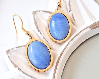 May include: A pair of gold-colored earrings featuring oval, light blue gemstones. The gemstones are set within a gold-colored frame and hang from gold-colored ear wires. The earrings are displayed on a white surface.
