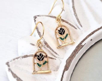 May include: Gold dangle earrings with a black rose inside a gold cage. The rose has green leaves.