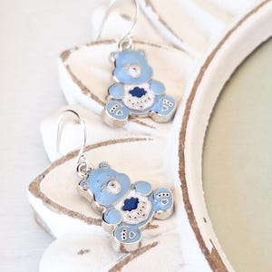 May include: A pair of silver dangle earrings featuring a blue Care Bear with a white heart and a blue cloud on its chest. The bear is sitting down with its paws in front of it. The earrings have a silver hook closure.