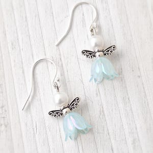 May include: A pair of angel-themed earrings. Each earring features a silver-colored metal angel with a pearl head and light blue flower-shaped body. The earrings hang from silver-colored ear wires. The background is a white, wooden surface.