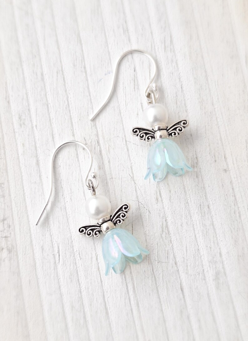 May include: A pair of angel-themed earrings. Each earring features a silver-colored metal angel with a pearl head and light blue flower-shaped body. The earrings hang from silver-colored ear wires. The background is a white, wooden surface.