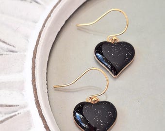 May include: A pair of gold dangle earrings with black heart-shaped charms. The charms have a sparkly, glittery finish.