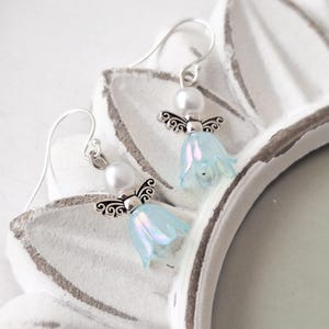 May include: A pair of dangle earrings featuring a light blue flower design with silver-colored angel wings and a white pearl. The earrings are displayed on a white, decorative surface. The earrings are a delicate and whimsical accessory.