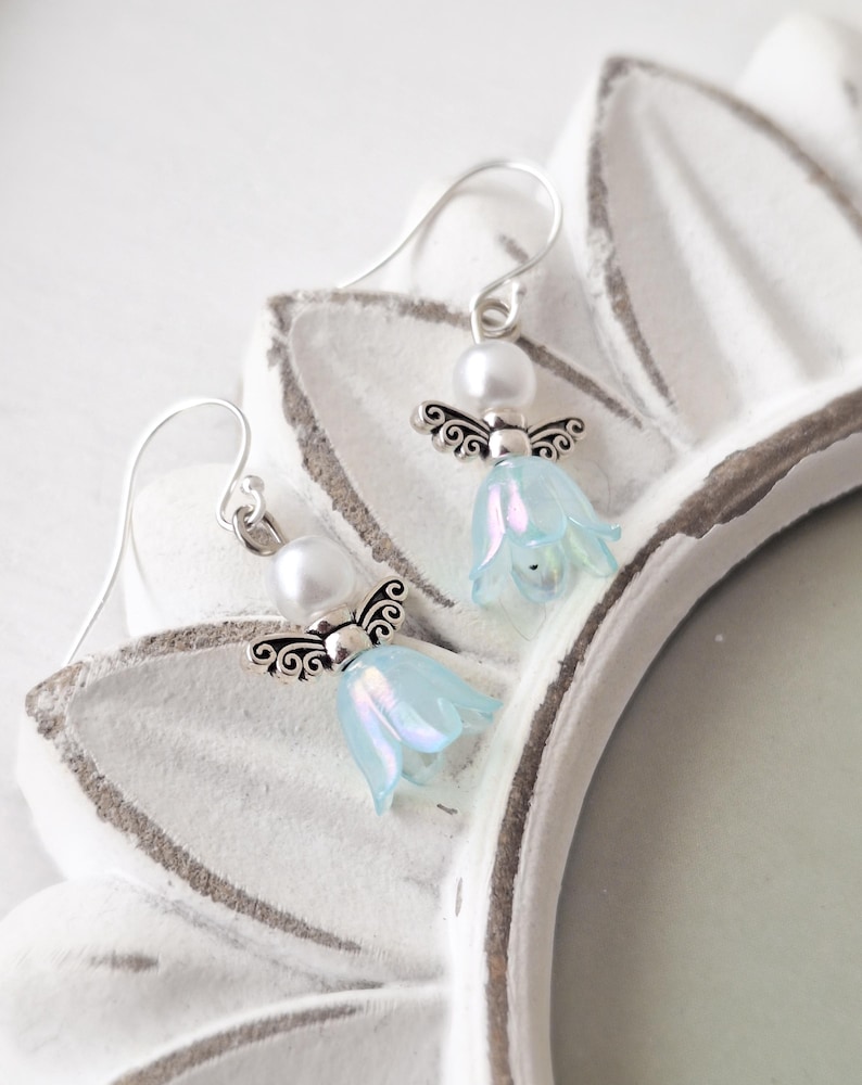 May include: A pair of dangle earrings featuring a light blue flower design with silver-colored angel wings and a white pearl. The earrings are displayed on a white, decorative surface. The earrings are a delicate and whimsical accessory.