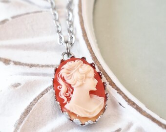 May include: Oval cameo pendant necklace featuring a profile of a woman with upswept hair in cream against a red background. The pendant is set in a silver-tone metal frame and hangs from a silver chain. A classic and elegant jewelry piece.