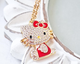 May include: A gold-tone metal necklace featuring a pendant shaped like a cat character. The pendant is encrusted with clear crystals and accented with red enamel on the bow and dress. The cat has black eyes and a yellow nose, hanging from a chain.