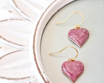 May include: A pair of gold heart-shaped earrings with a pink glitter finish.