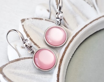 May include: A pair of dangle earrings with a light pink, circular stone set in a silver-colored metal frame. Each earring has a small white pearl accent near the hook. The earrings are displayed on a white, decorative surface.