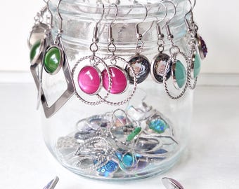 May include: A collection of assorted earrings with silver-toned hooks and settings, featuring various colorful cabochons and patterned designs, displayed hanging from the rim of a clear glass jar with additional loose earrings placed in front.