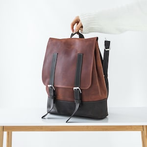 以下が含まれることがあります： ダークグレーのベースと、シルバーの金属製バックルが付いた2本の垂直なダークグレーのストラップが特徴のブラウンのレザーバックパック。フラップ式の開閉部と調節可能なショルダーストラップを備えたツートンカラーのデザインです。