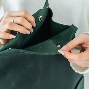 May include: A dark green leather bag with a flap closure. The bag is being opened, revealing the interior. The bag has a simple design with a snap closure. The bag is held by a person wearing a white jumper.