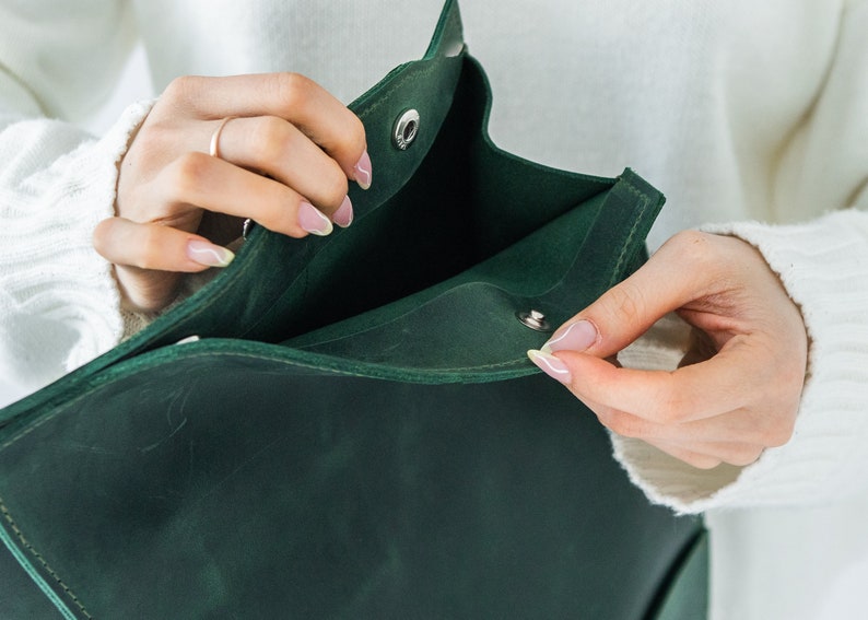 May include: A dark green leather bag with a flap closure. The bag is being opened, revealing the interior. The bag has a simple design with a snap closure. The bag is held by a person wearing a white jumper.