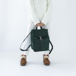 May include: A dark green suede backpack with a flap closure and silver-tone hardware. The backpack has adjustable black straps and a top handle. The bag is held by a person wearing a white sweater and trousers. The person is also wearing brown boots.